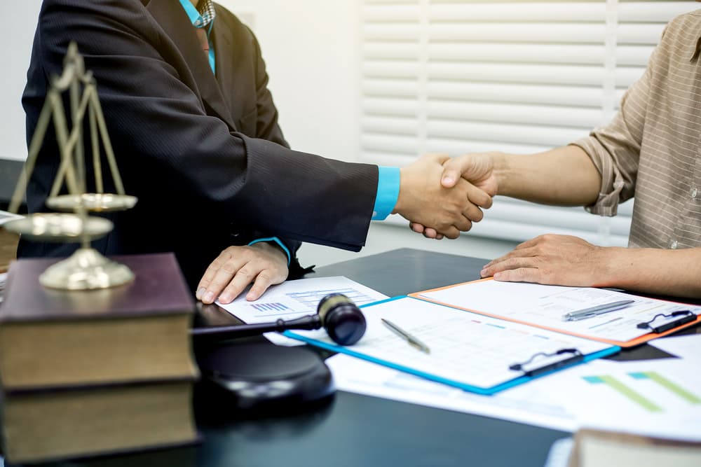 Two businessmen shaking hands in agreement with lawyers discussing contract in background.