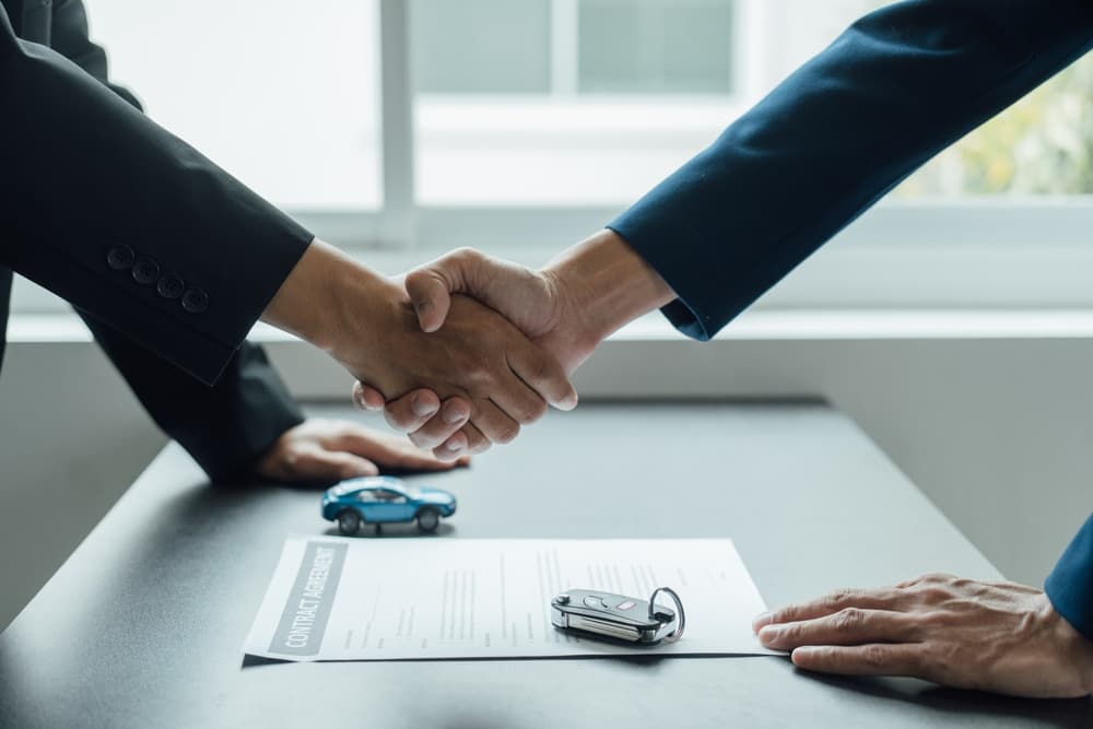Two individuals shaking hands over a contract with a model car nearby.